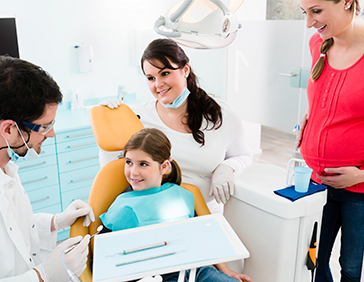 Niña recibiendo atención dental en una clínica mientras un adulto la acompaña y otro profesional sanitario la atiende.