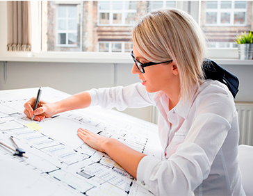 Persona trabajando sobre planos técnicos en una mesa de dibujo dentro de una oficina luminosa.