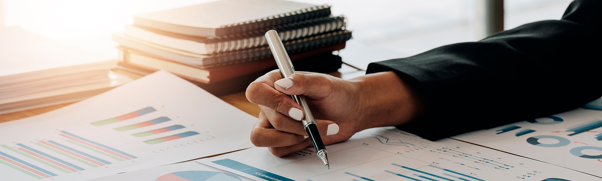 Imagen de una persona escribiendo con un bolígrafo junto a una mesa con gráficos y documentos de gestión.
