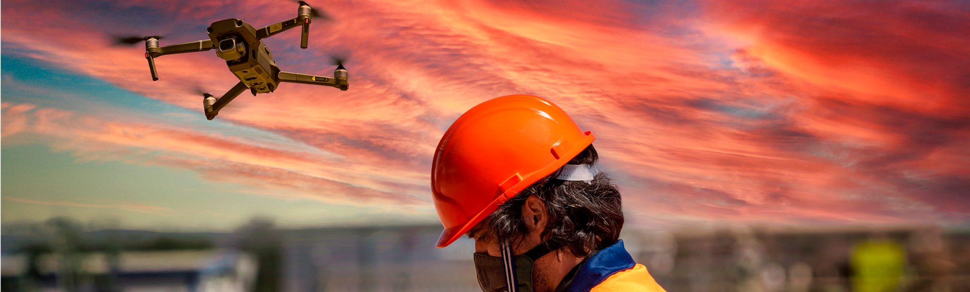Imagen de un dron en pleno vuelo al atardecer mientras una persona con casco de seguridad maneja un control remoto